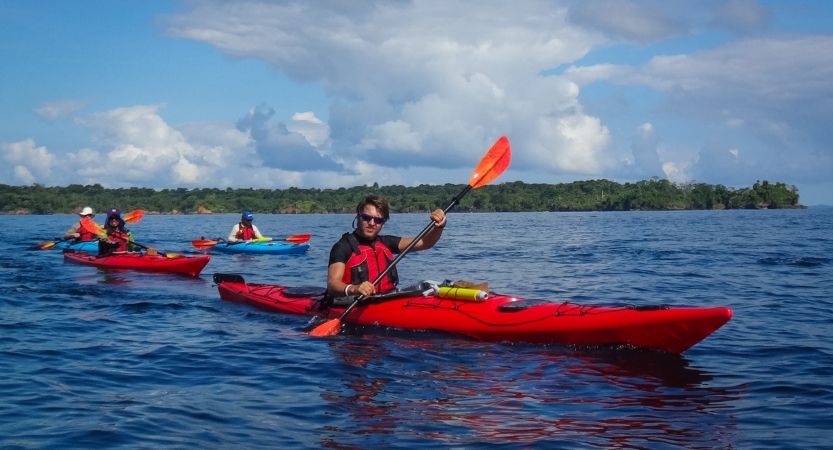 Four students paddle four kayaks on calm blue water. There is a tree-lined shore in the background.